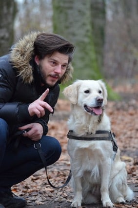 A bearded man wearing a parka, crouched down next to a sitting golden retriever and holding the dog's leadh. The man is giving a hand cue to the dog. The dog is looking at something out of frame and appears to be waiting expectantly.