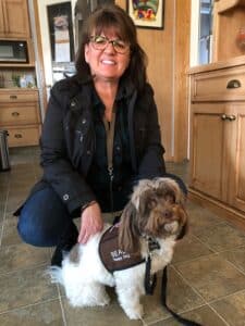 Beau, a brown and white Havanese therapy dog, wearing a Pet Partners vest, with his handler Judy behind him.