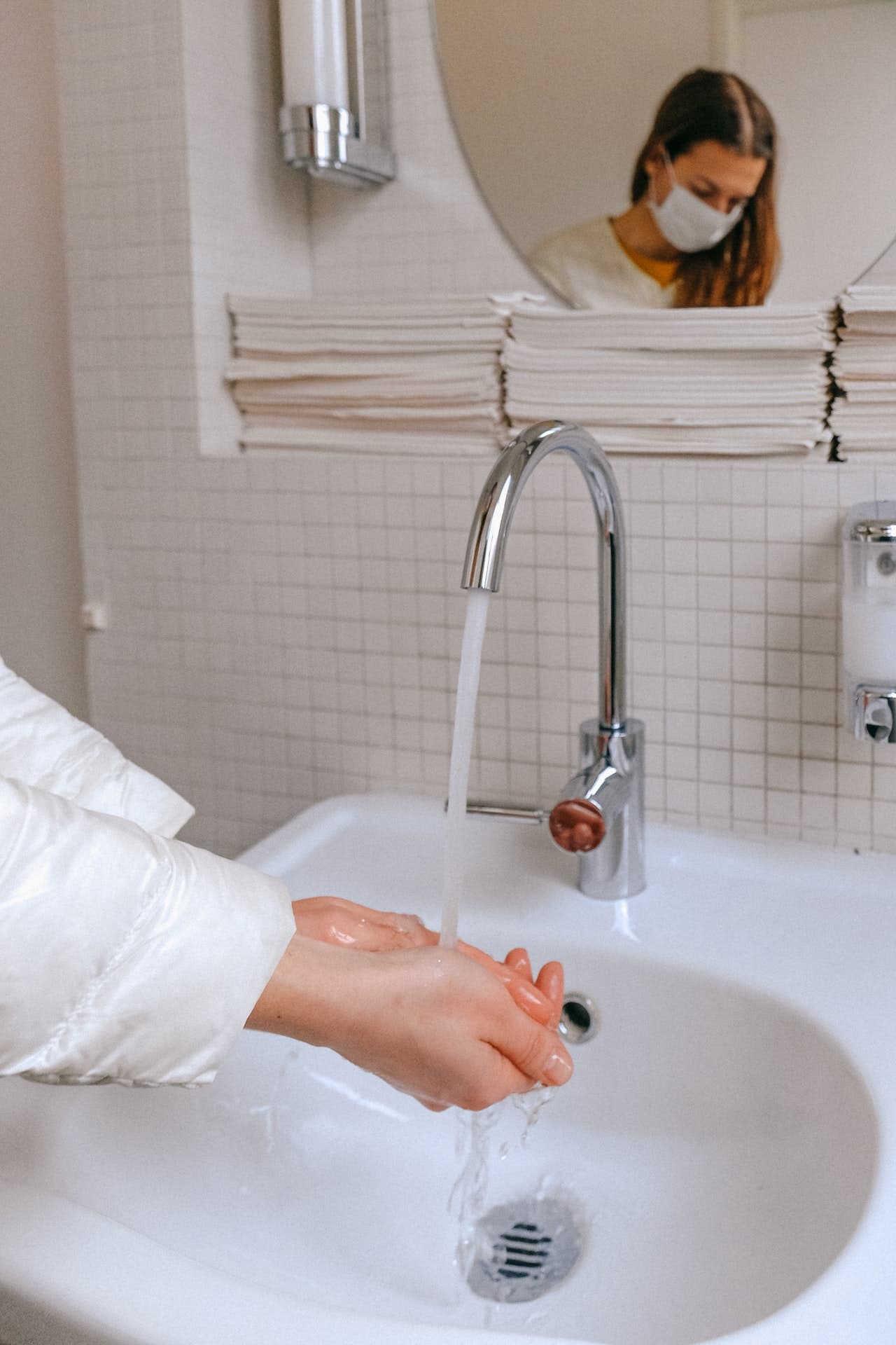 A woman washing her hands; her masked face is shown in a mirror above the sink. Photo by Anna Shvets from Pexels