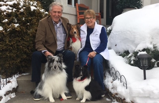 A smiling man and woman sit in front of a house with three Shetland sheepdogs