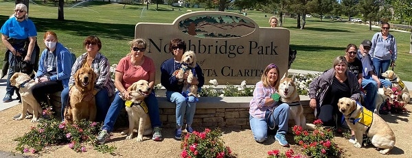 Guide dogs in training and their handlers pose with several Pet Partners therapy dog teams in front of a park sign after a walk. 