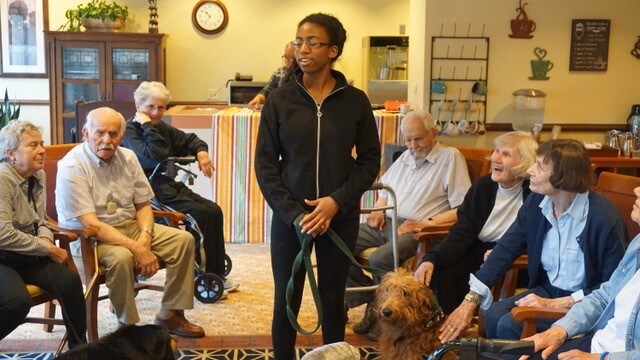 A woman with a dog stands in the middle of a group of people at a nursing home.
