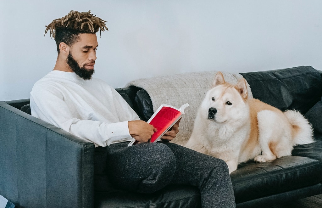 A man sitting on a couch reading a book with a dog watching him. Photo by Zen Chung from Pexels.