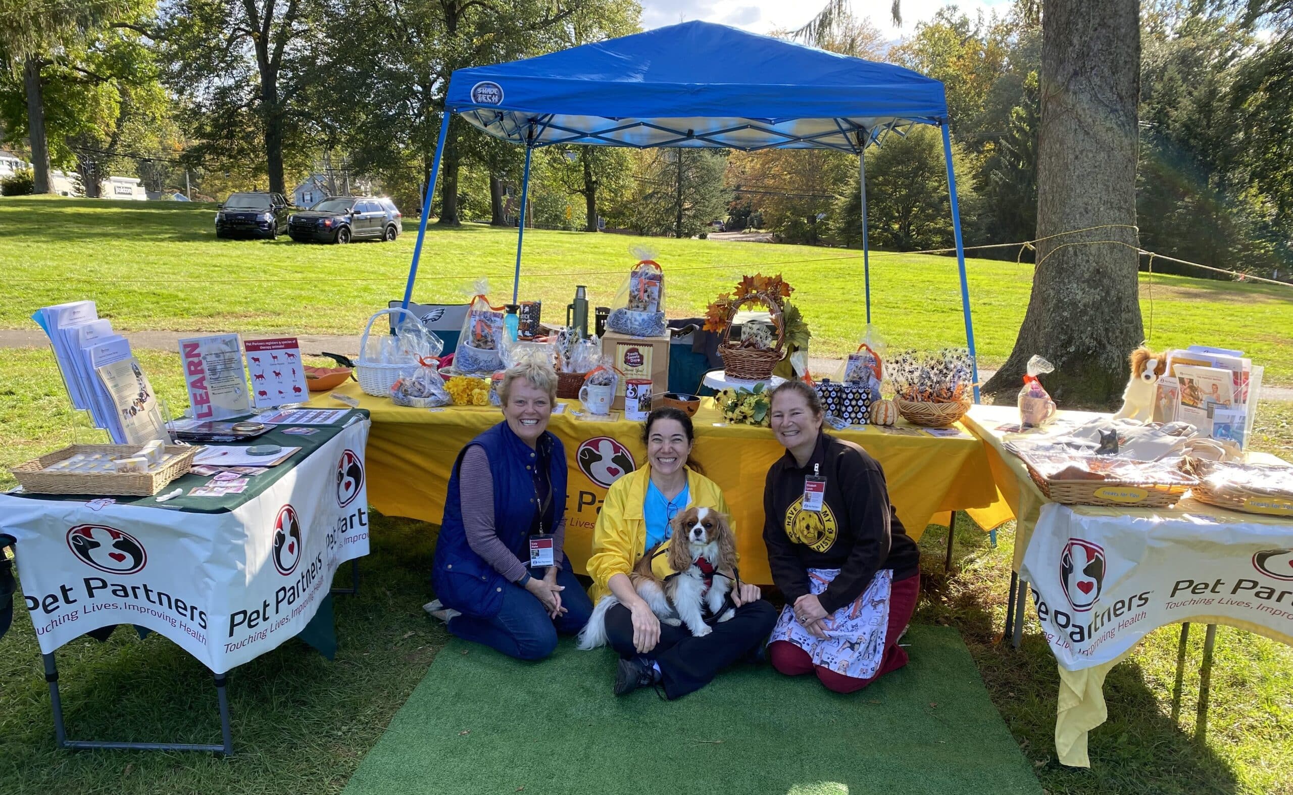 Three Pet Partners volunteers, smiling, sit on the ground in front of the Pet Partners booth at the Westport Dog Festival.