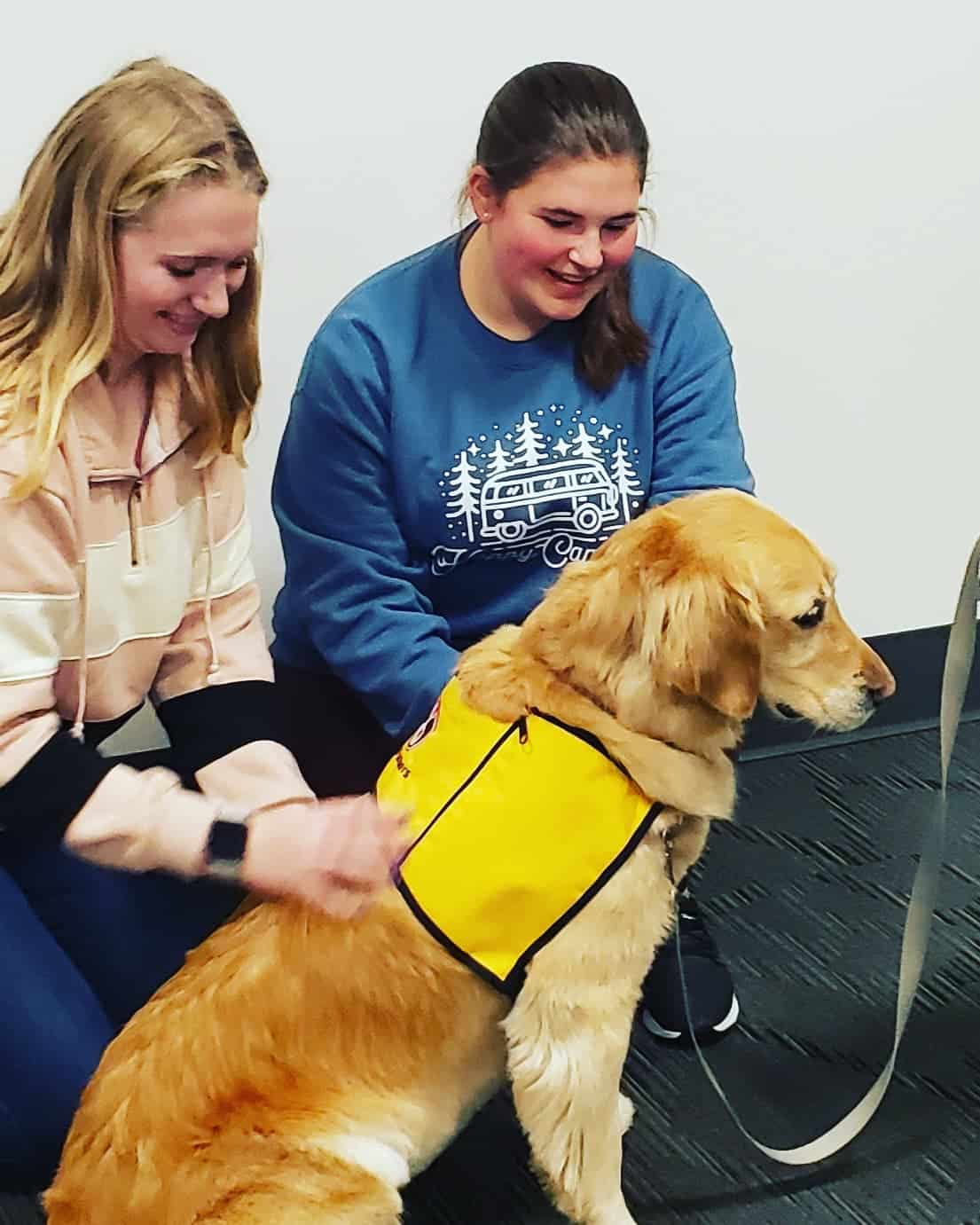 Two young women smile as they pet a golden retriever therapy dog wearing a Pet Partners vest