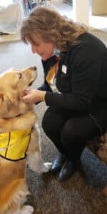 A sitting golden retriever therapy dog wearing a Pet Partners vest looks up at a smiling woman who is crouched down to pet her