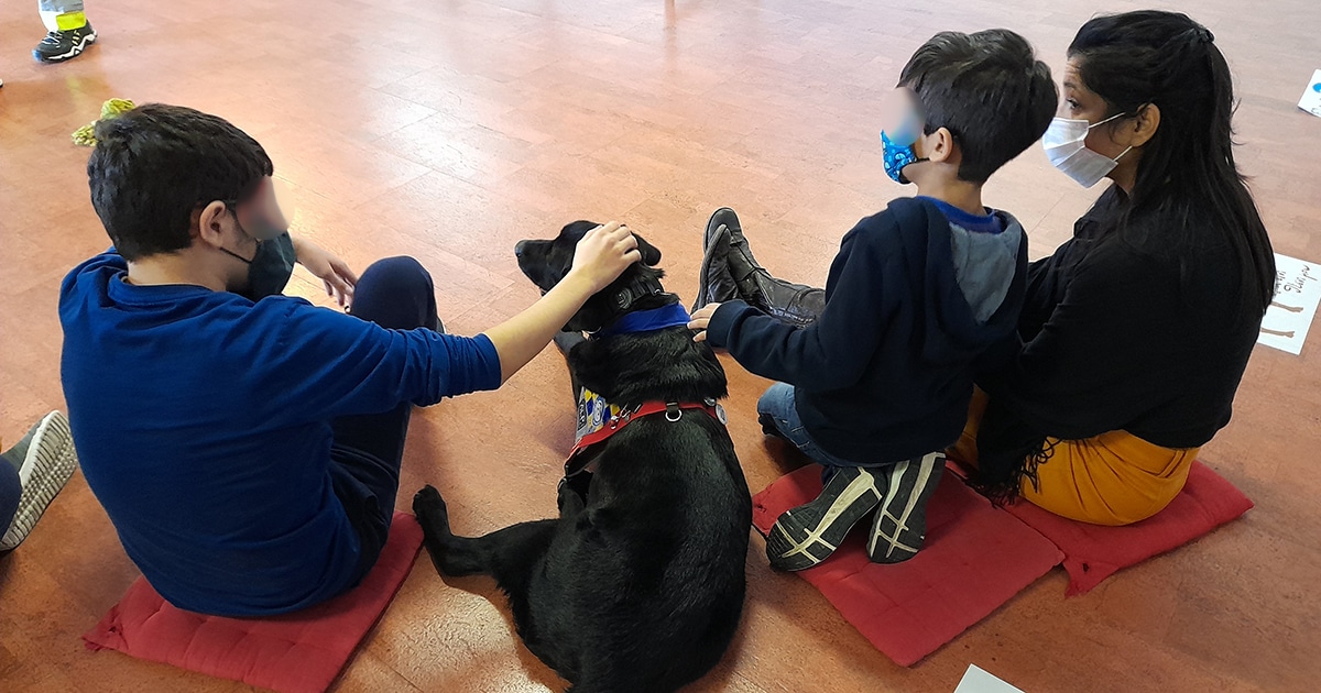 A black Labrador retriever therapy dog lies on the floor between two children and an adult, all masked.