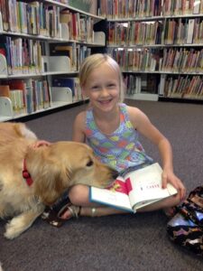 A smiling girl holds an open book on her lap, with a golden retriever next to her lying with her head on the book.