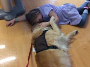 A child wearing a hospital gown lies on the floor, forehead to forehead with a golden retriever therapy dog.