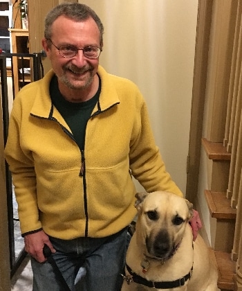 A therapy dog handler wearing glasses and a yellow jacket stands next to his mixed-breed therapy dog, with one hand on her back and her leash in his other hand.