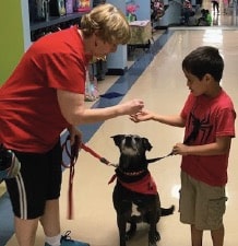 A therapy dog handler guides a student in how to give a treat to her therapy dog as they stand in a school hallway.