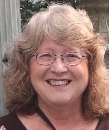A woman with wavy blond hair and glasses smiles at the camera.