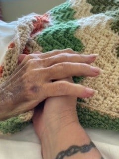 A handler holds the hand of a hospice client on a patterned blanket.