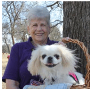 A smiling woman holding a basket containing a blonde Pekingese mix therapy dog.