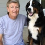 A man wearing a gray shirt sits with an arm around his Bernese mountain dog therapy dog.