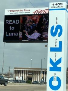 An electronic sign for the Central Kansas Library System shows a photo of a Bernese mountain dog therapy dog with a child reading to her and the words "READ to Luna"