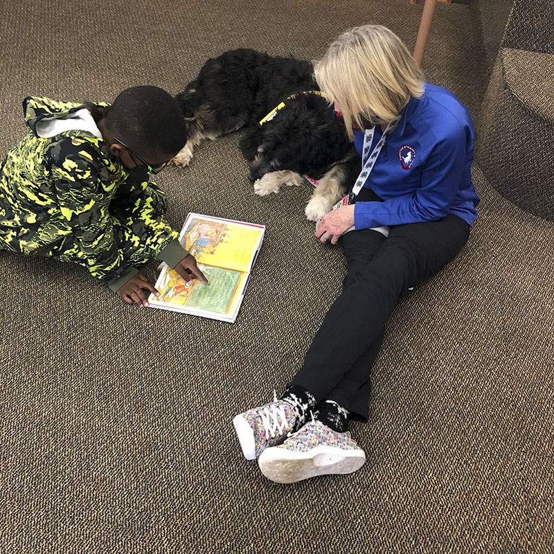 Therapy dog handler participates in Read With Me in a school, while a young boy reads a book to her dog.