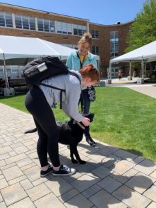 A high school student pets a black Lab therapy dog while the handler watches and supervises.