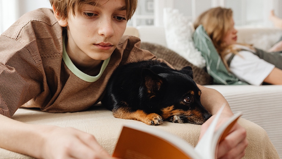 Boy reads a book while his dog sits by his side.