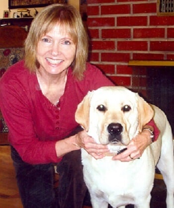 A smiling woman embraces her yellow Lab therapy dog, both looking at the camera.