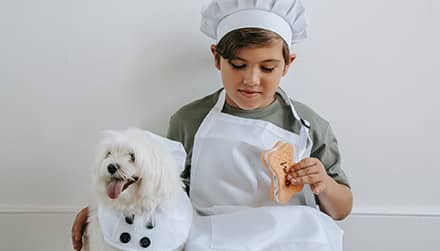 Boy dressed like a chef holds a cookie while sitting next to a white dog wearing an apron.