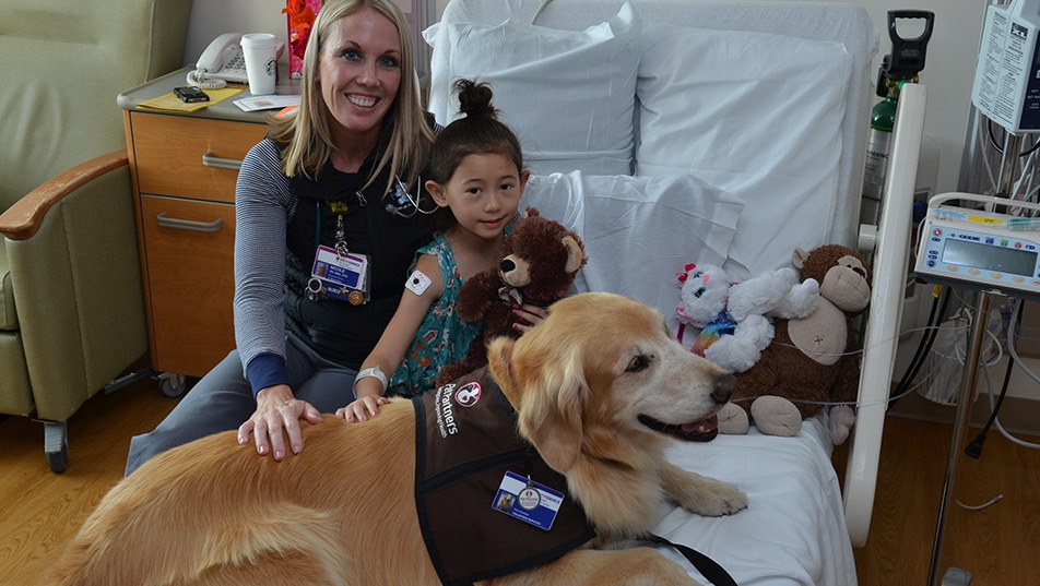 A Pet Partners therapy animal handler and her dog sit on a bed with a young patient they are visiting.