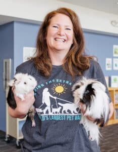 A smiling woman with long hair, wearing a National Therapy Animal Day T-shirt, holding a shorthaired white guinea pig in her right arm and a long haired black & white guinea pig in her left arm.