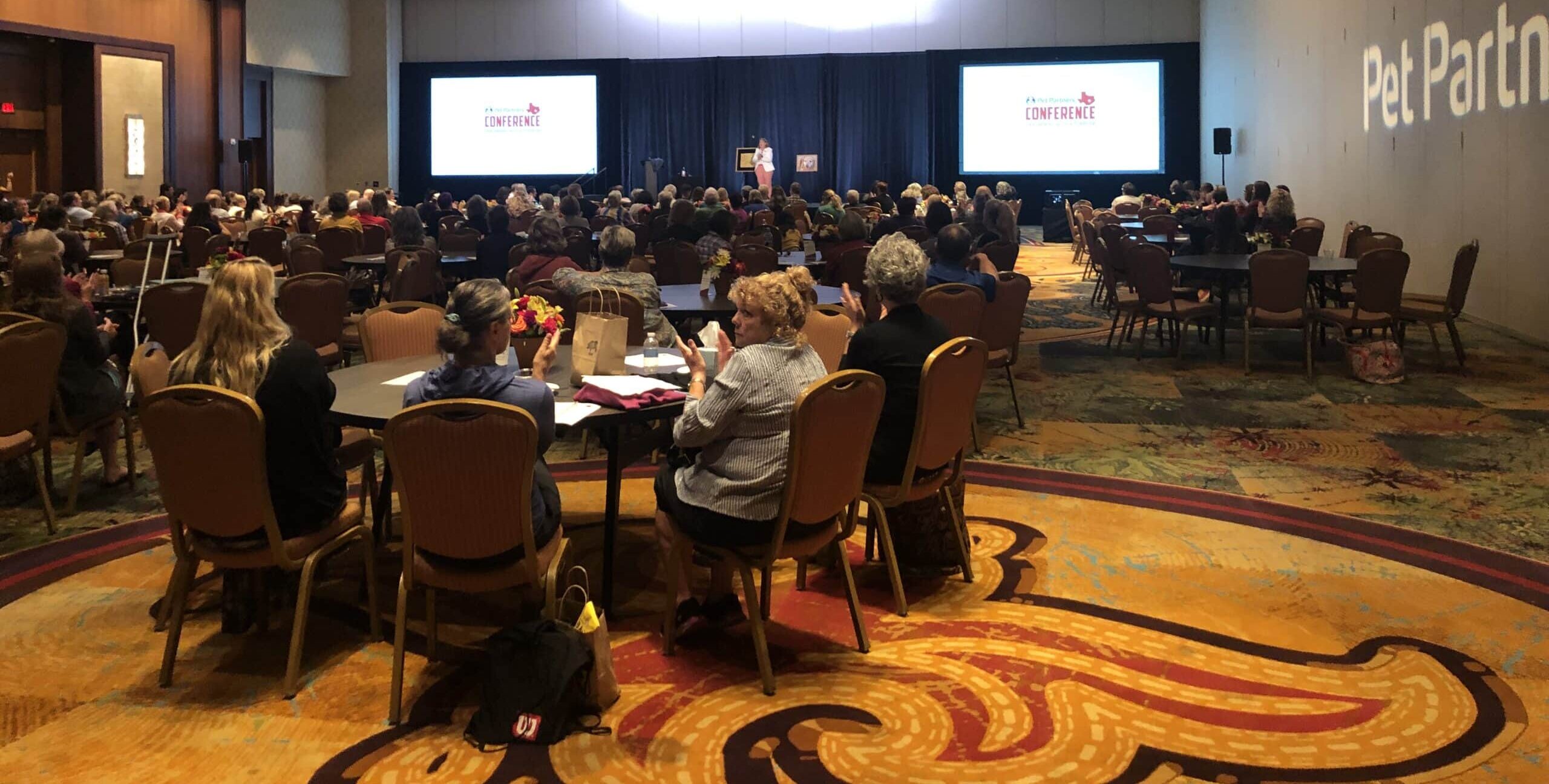 A ballroom full of people at tables during a gala dinner, with screens for the Pet Partners 2019 conference at the front of the room.