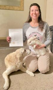 A therapy animal handler holds a certificate and poses with her therapy dog and rabbit.