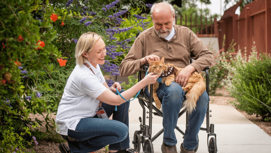 A woman crouches next to a man in a wheelchair holding a therapy cat