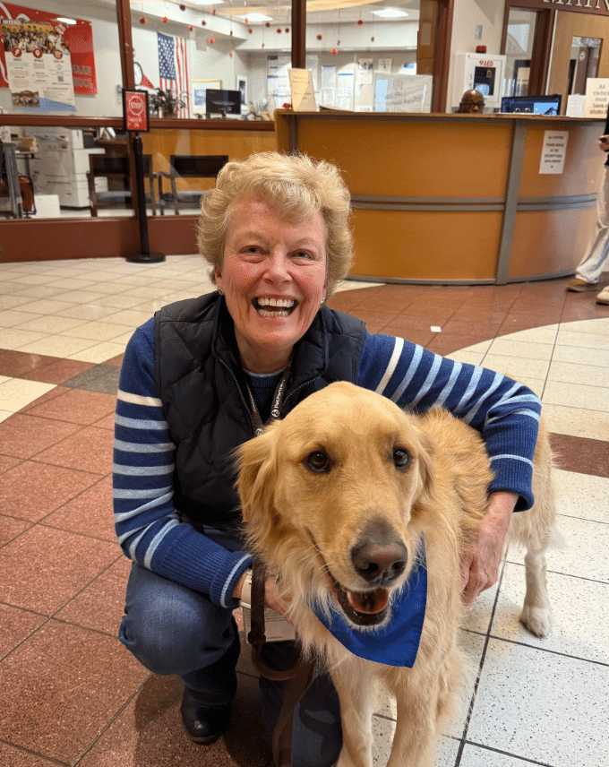 Kathy Rahe smiling with River, a Golden Retriever therapy dog, wearing a blue bandana inside a community facility.
