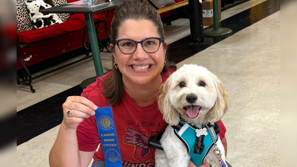 Smiling woman kneels beside her happy white dog, proudly holding an AKC Canine Good Citizen ribbon after passing the CGC test.